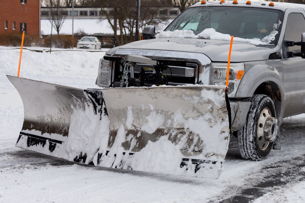 A snow plow truck clearing snow from a road, with snow buildup on the front blade and snowy surroundings visible.